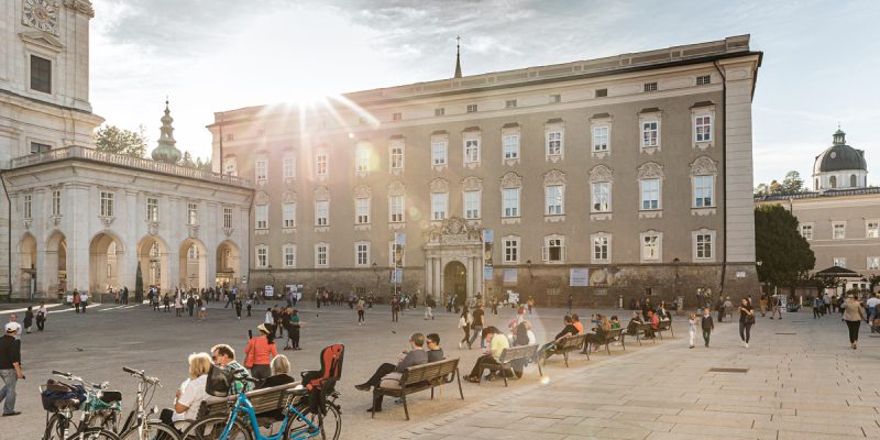 Residenzplatz mit vielen Menschen auf den Bänken und gehend über den Platz, im Hintergrund die barocke Fassade der Alten Residenz mit Hauptportal und Terrasse mit Übergang in den Dom. Foto im Gegenlicht der Sonne am Nachmittag aufgenommen.