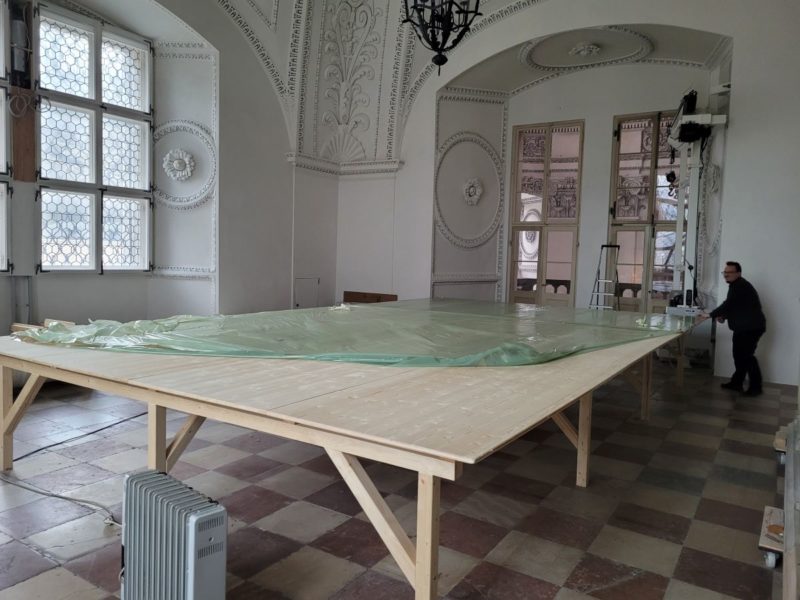 Arbeitsraum mit großem Tisch in der Mitte. Dieser hat eine ca. 30m² große Arbeitsfläche mit Schutzfolie die gerade aufgelegt wird.